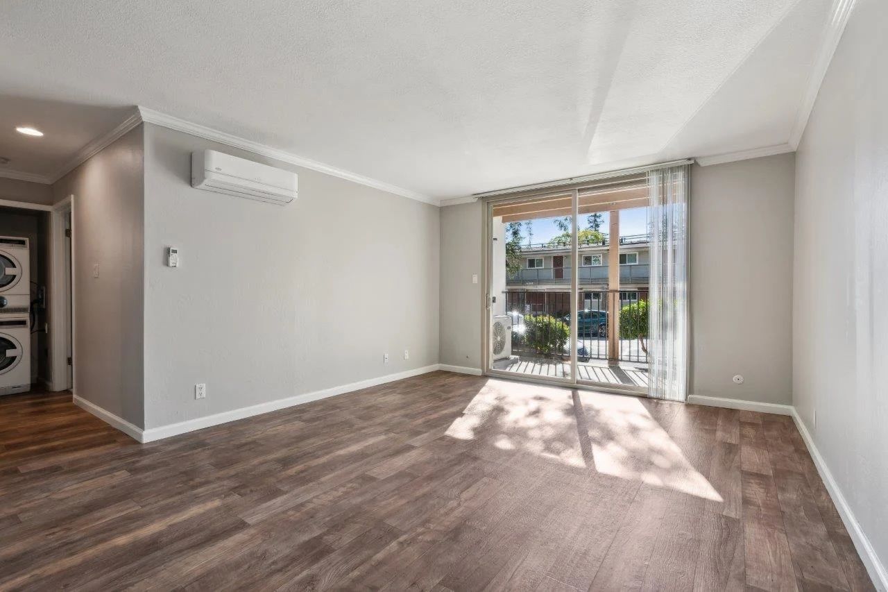 Empty living room with wooden floors, sliding glass door, air conditioning unit, and washer/dryer.