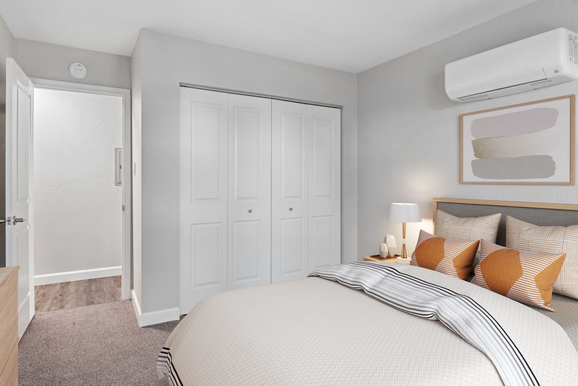 Bright bedroom with white closet doors, patterned bedding, wall art, and a wall-mounted air conditioner.
