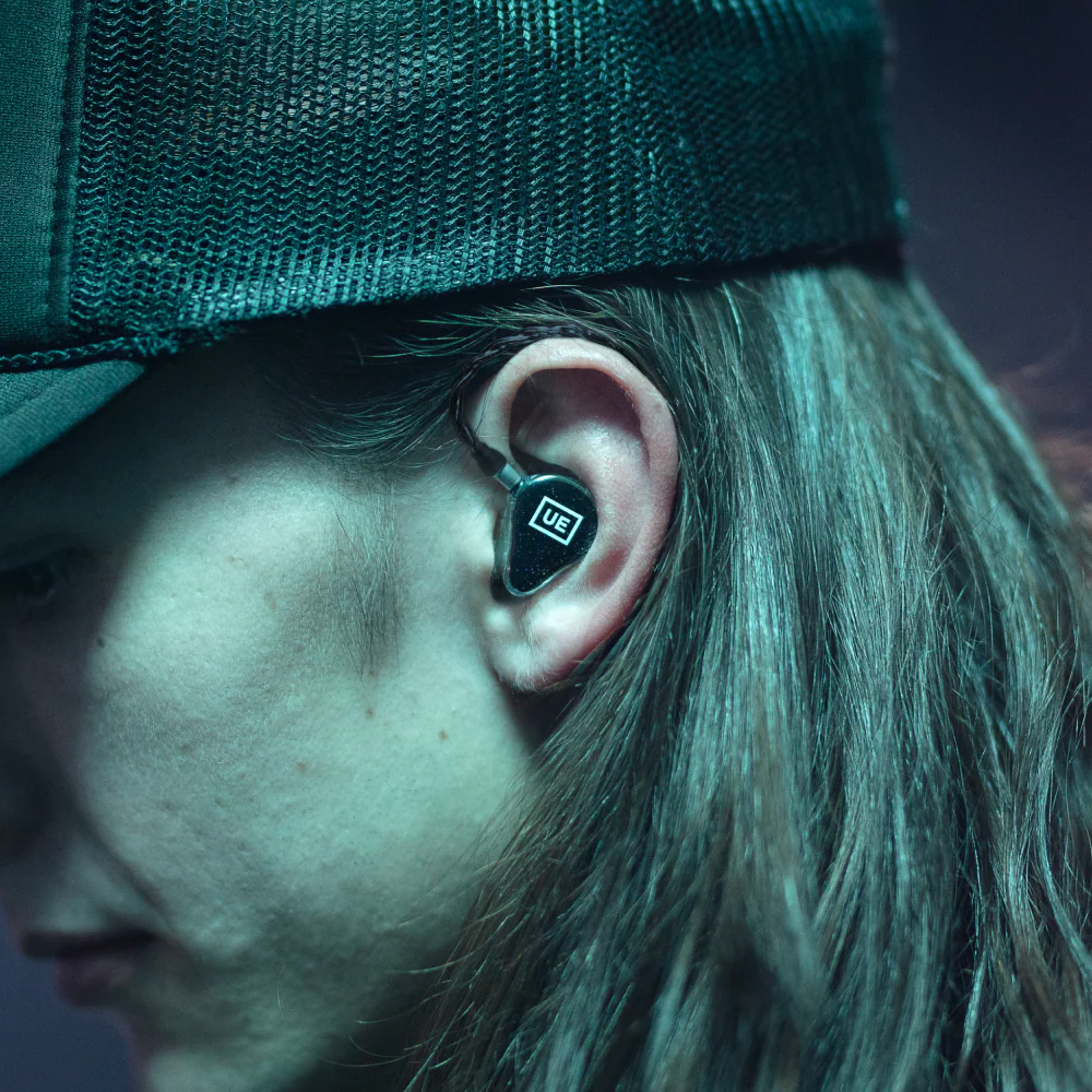 Person wearing a black earbud in their ear, under a black cap, in a dark setting.
