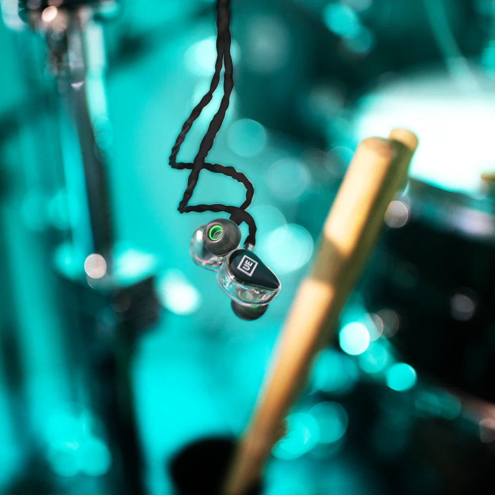 Earphones hanging in front of a drum, with a drumstick in the foreground. Teal and gold tones.