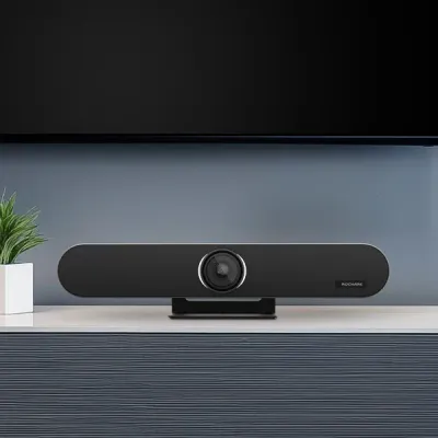 A black video conferencing soundbar on a shelf below a TV screen. A small plant is to the left.