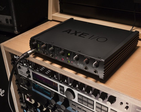 Black Axe I/O audio interface on a wooden shelf, connected to a cable. Other audio equipment is below.