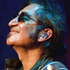 Man with sunglasses and in-ear monitors, looking up, smiling. Dark hair, blue lighting.