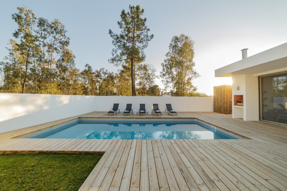 Swimming pool with wooden deck and lounge chairs, next to white building and green lawn.