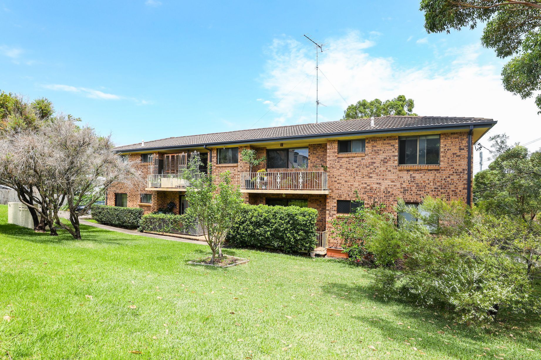 Two-story brick building with balconies, surrounded by green lawn and trees under a blue sky.