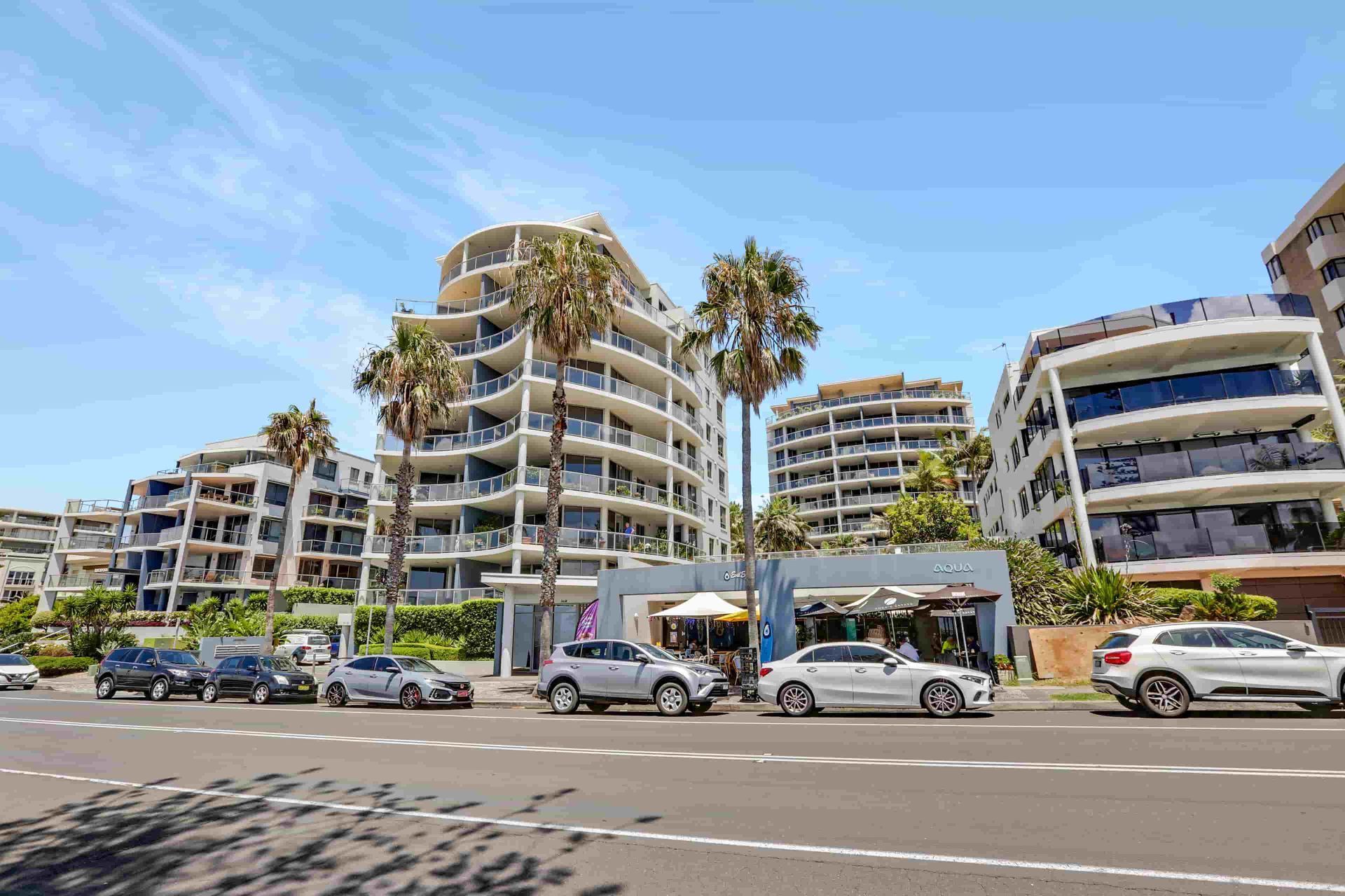 Apartment buildings and parked cars line a street with palm trees under a blue sky.