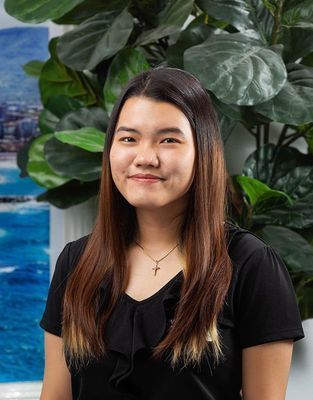 Young Asian woman with long brown hair smiles in front of a green plant and seascape background.