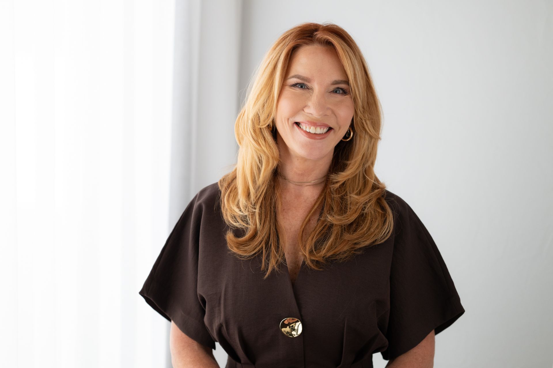 Woman with blonde hair smiles, wearing a dark blazer over a patterned blouse, standing on a white background.