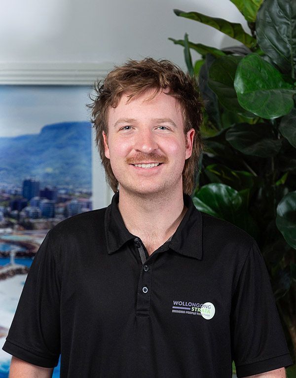 Man with a mullet and mustache smiling in a black polo shirt, next to greenery and a framed picture.