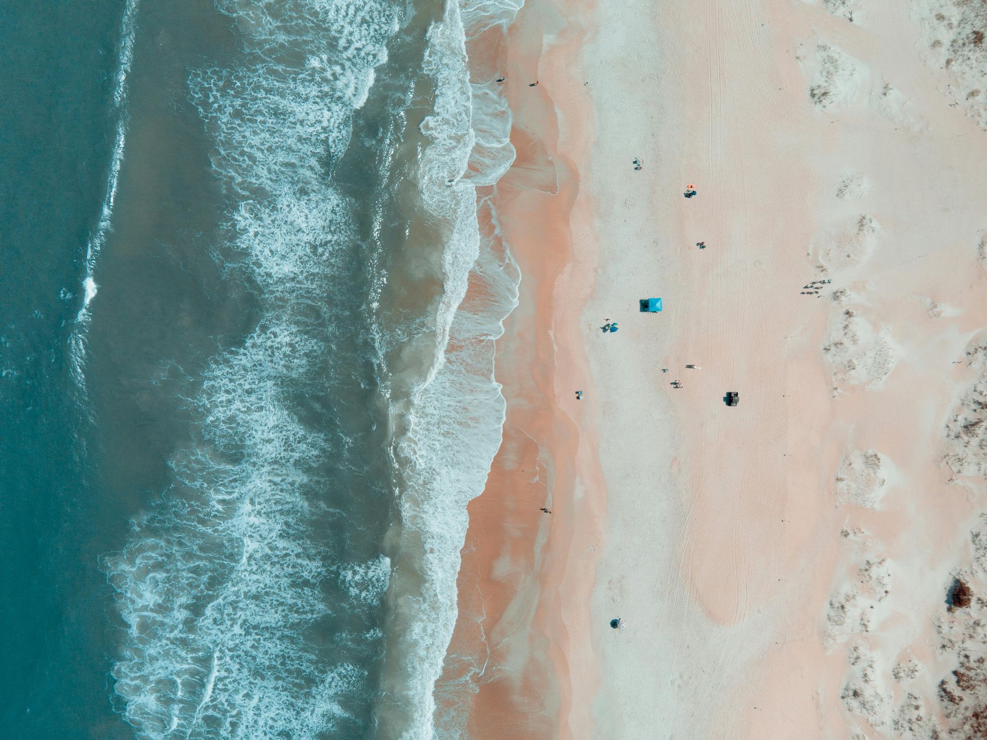 Aerial view of turquoise waves washing onto a pale sandy beach with a few tiny beachgoers and umbrellas