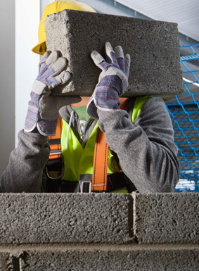 Obrero de la construcción con casco y chaleco de seguridad, cargando un bloque de hormigón sobre la cabeza.