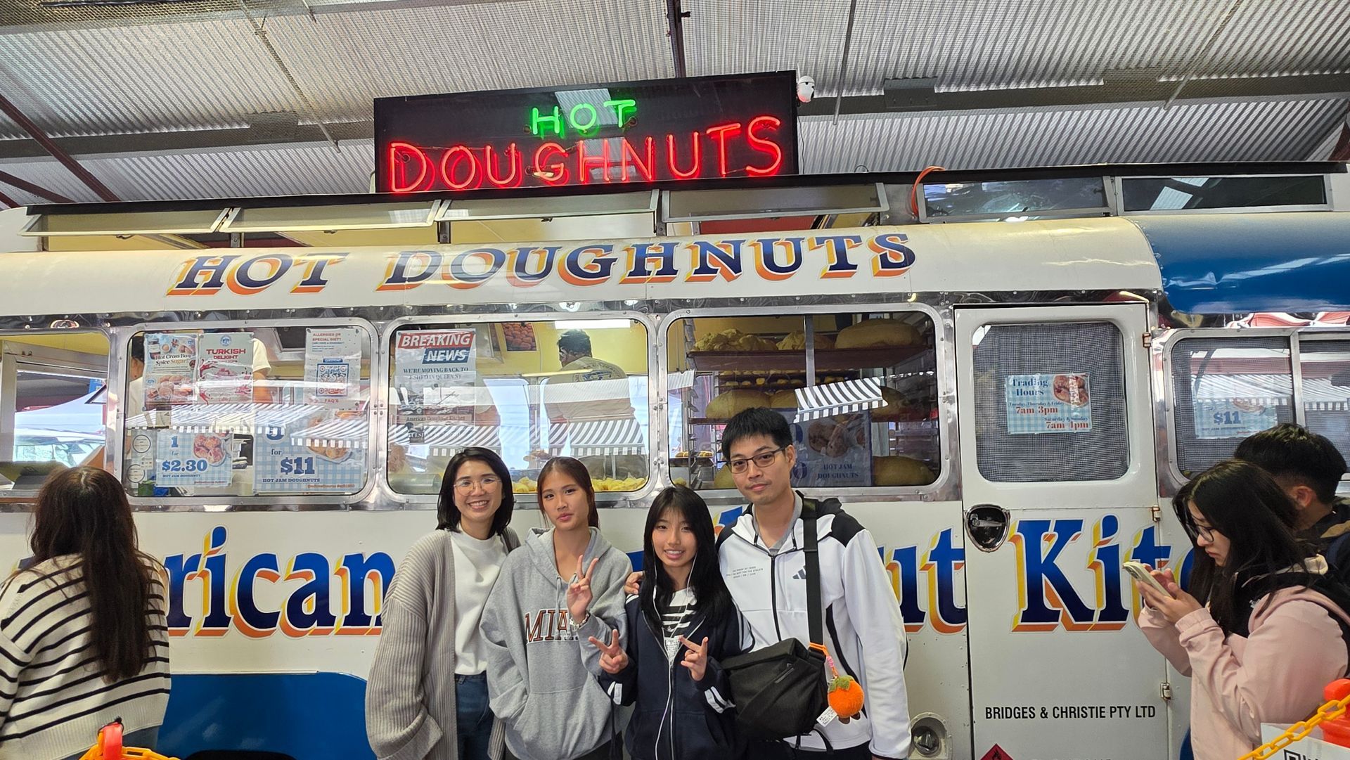 People posing in front of a Hot Doughnuts stand with a neon sign and displayed pastries