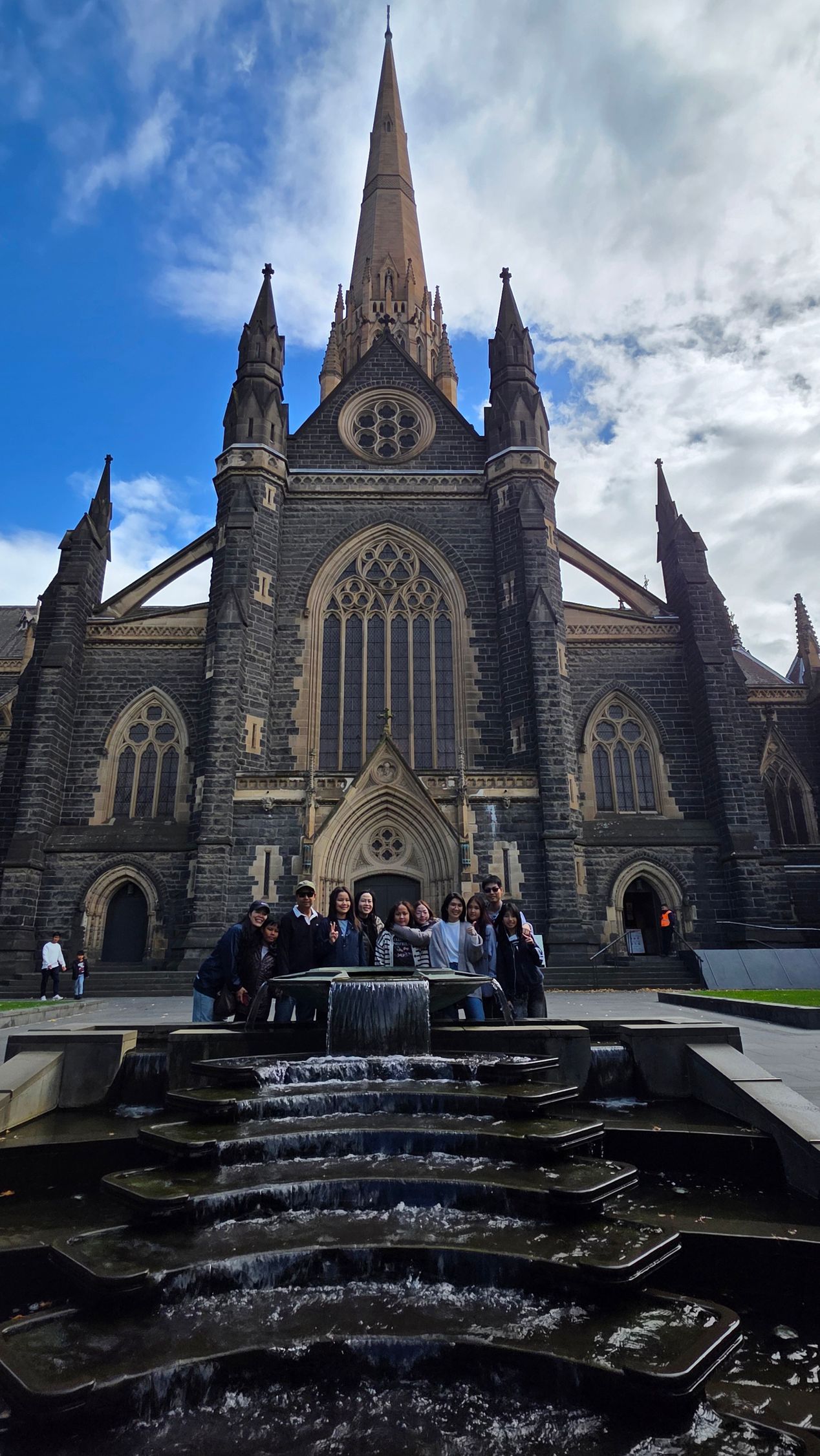 Gothic cathedral with a tall spire and a fountain in front under a cloudy sky