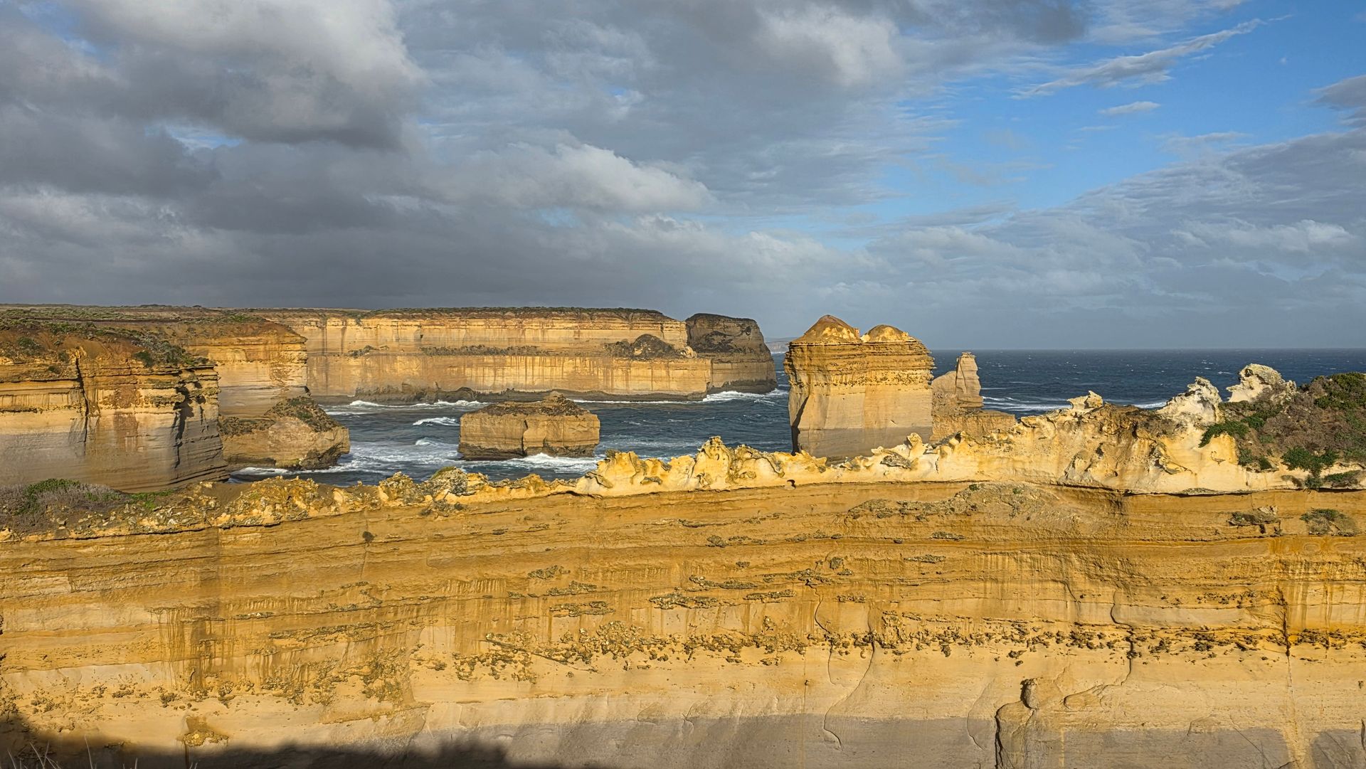 Sunlit limestone cliffs above a calm blue sea under a cloudy sky