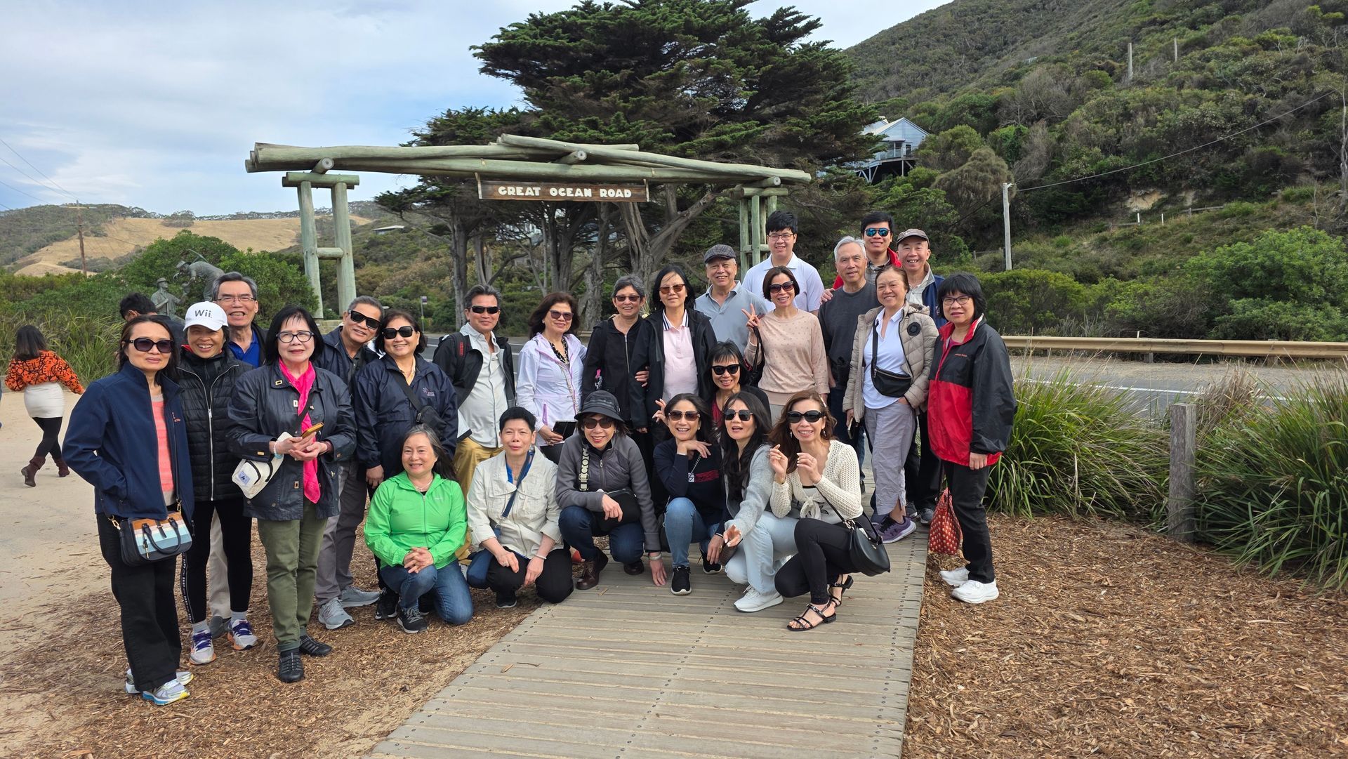Large group posing outdoors on a path under a pergola, with greenery and hills in the background.