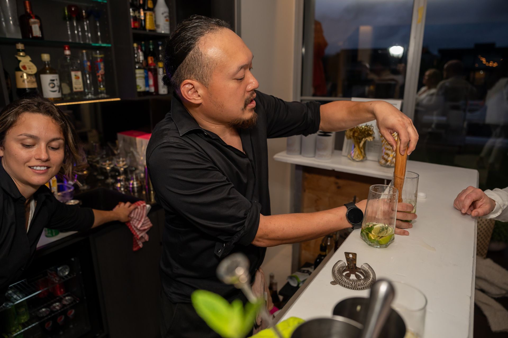 Bartender pouring a drink at a bar while a woman smiles nearby and another hand reaches in from the right