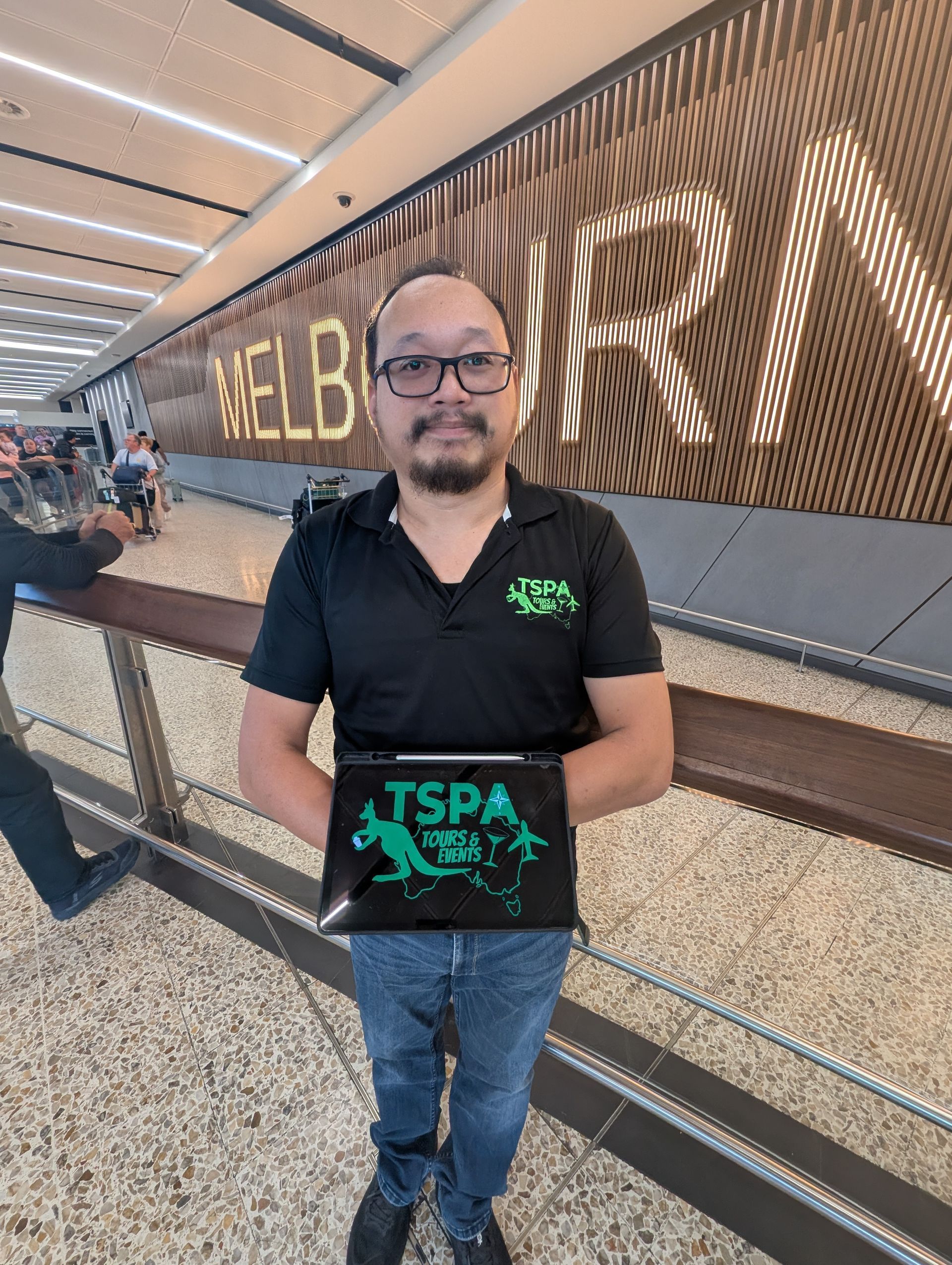 Man in black polo holding a green sign in a bright airport terminal with a wood slat wall