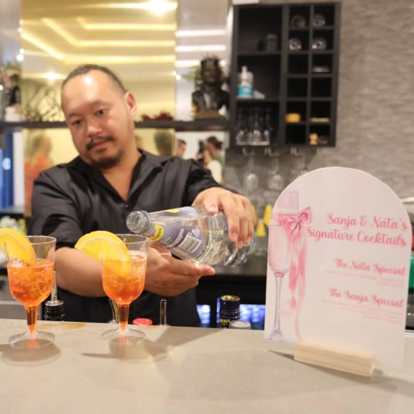 Bartender pouring liquor into orange cocktails beside a wedding sign at a bar