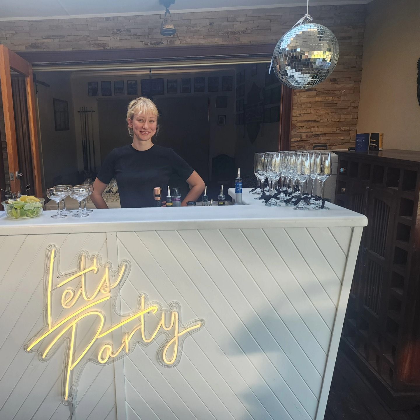 Outdoor bar with a woman behind a white counter, neon “Let’s Party” sign, and disco ball decorations