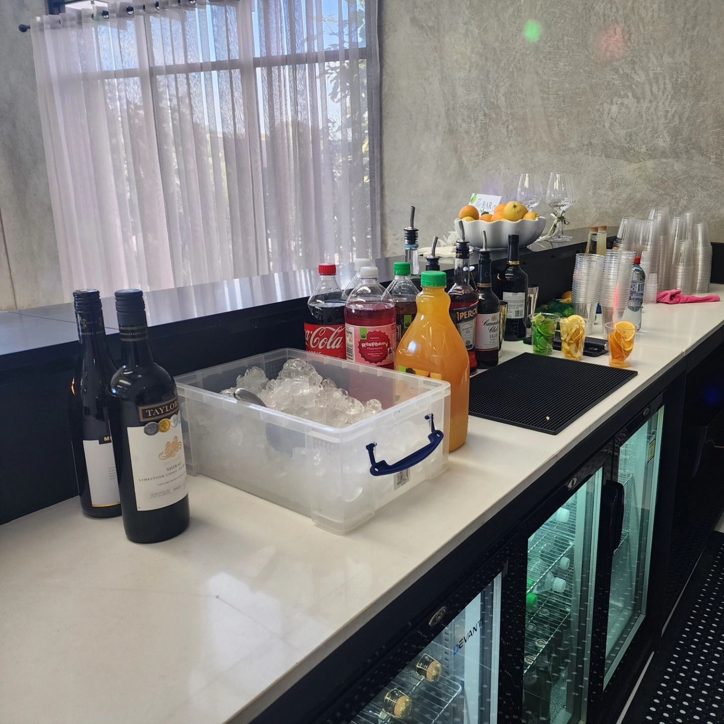 Bar counter with bottles, mixers, and an ice bin, set against a window with sheer curtains.