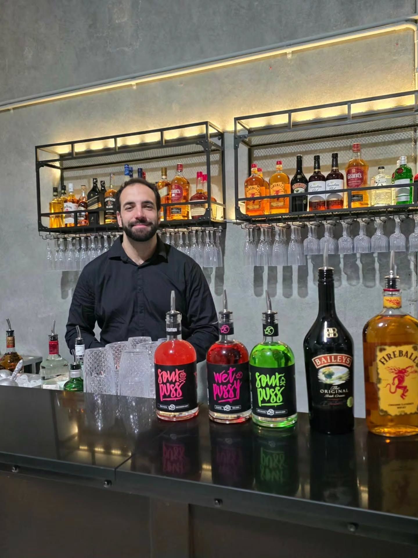 Bartender behind a bar with colorful liquor bottles and hanging glasses on shelves.
