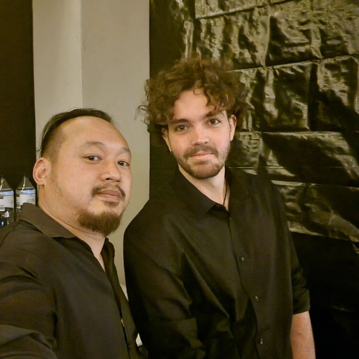 Two people in black shirts posing indoors against a textured dark wall.