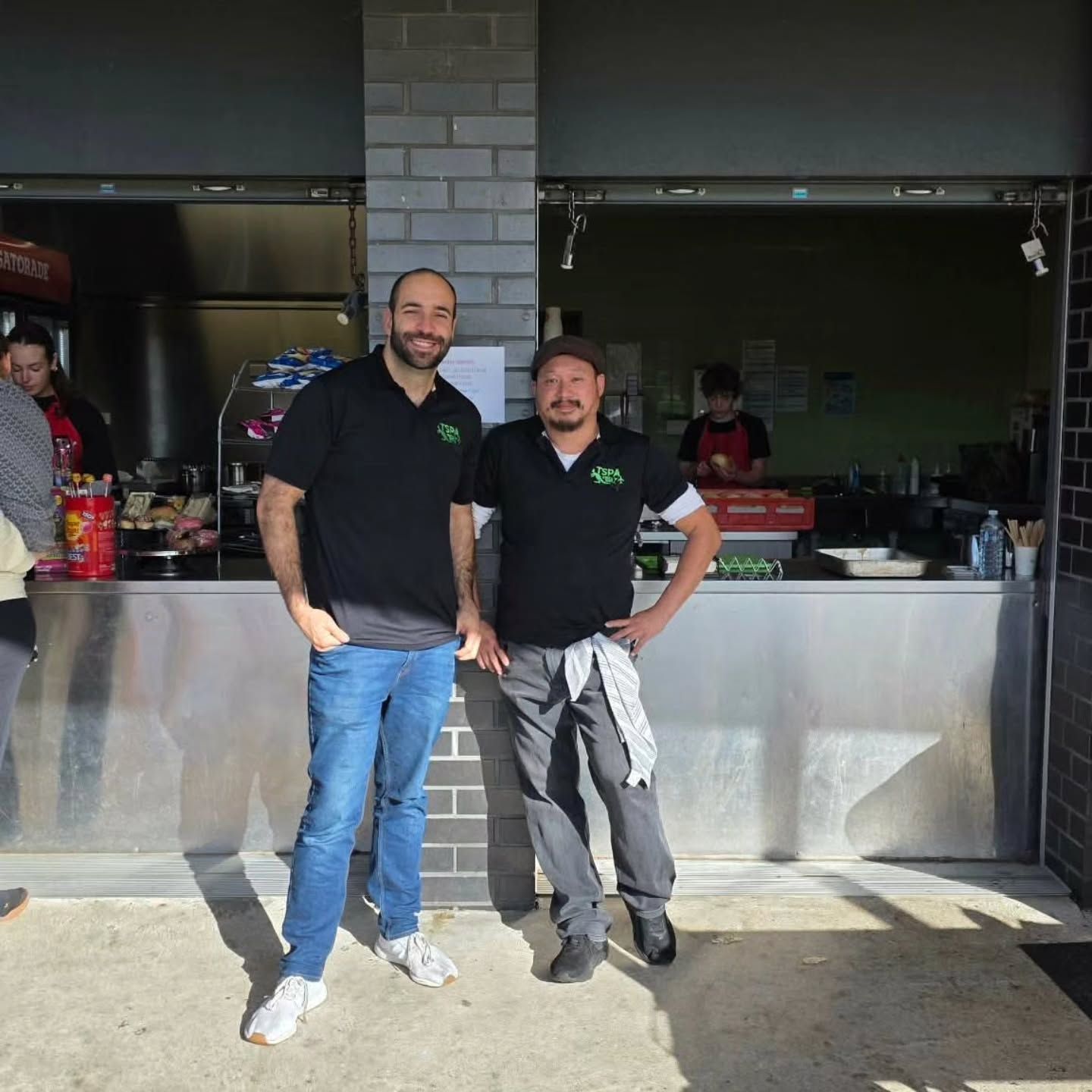 Two smiling restaurant staff in black shirts stand behind a metal counter.