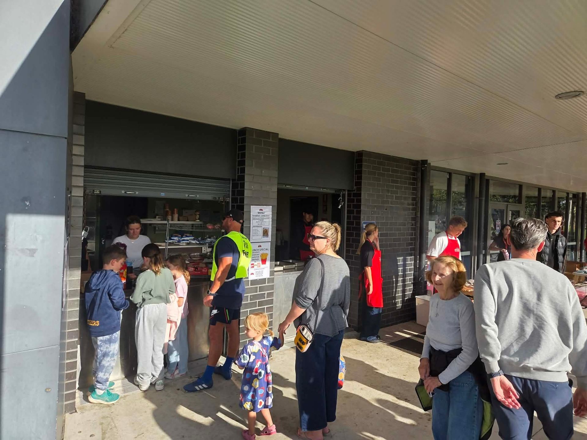 People gathered outside a building entrance under an overhang, with a worker in a reflective vest near the door.