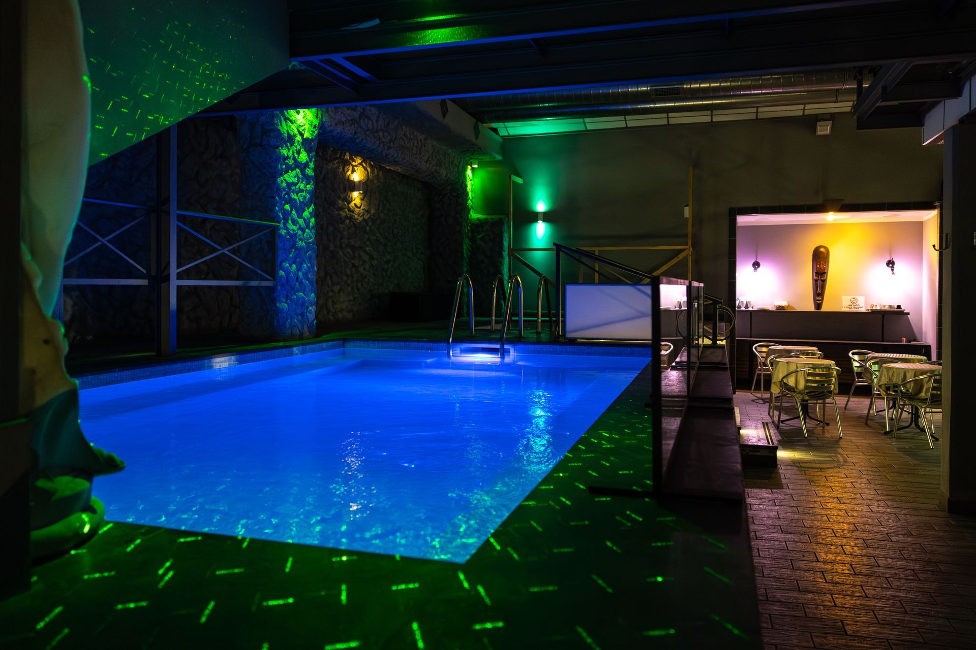 Piscina coperta con acqua blu, luci verdi e una zona lounge illuminata da una luce calda adiacente.