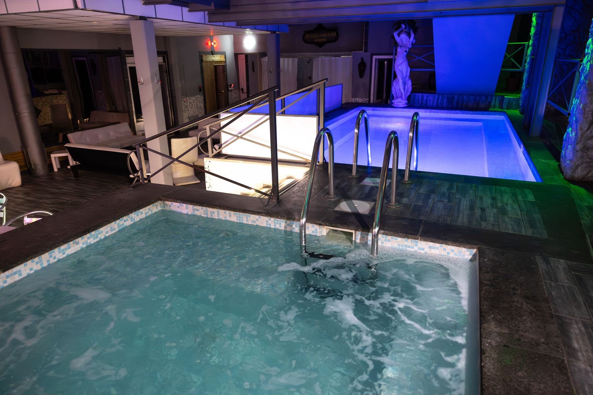 Area piscina e vasca idromassaggio illuminata con ringhiere, luci blu e una piccola statua sullo sfondo.