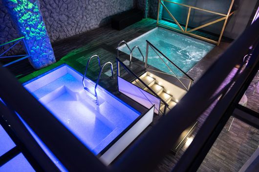 Due vasche idromassaggio luminose accanto a una piscina coperta con ringhiere in metallo e pareti in pietra.