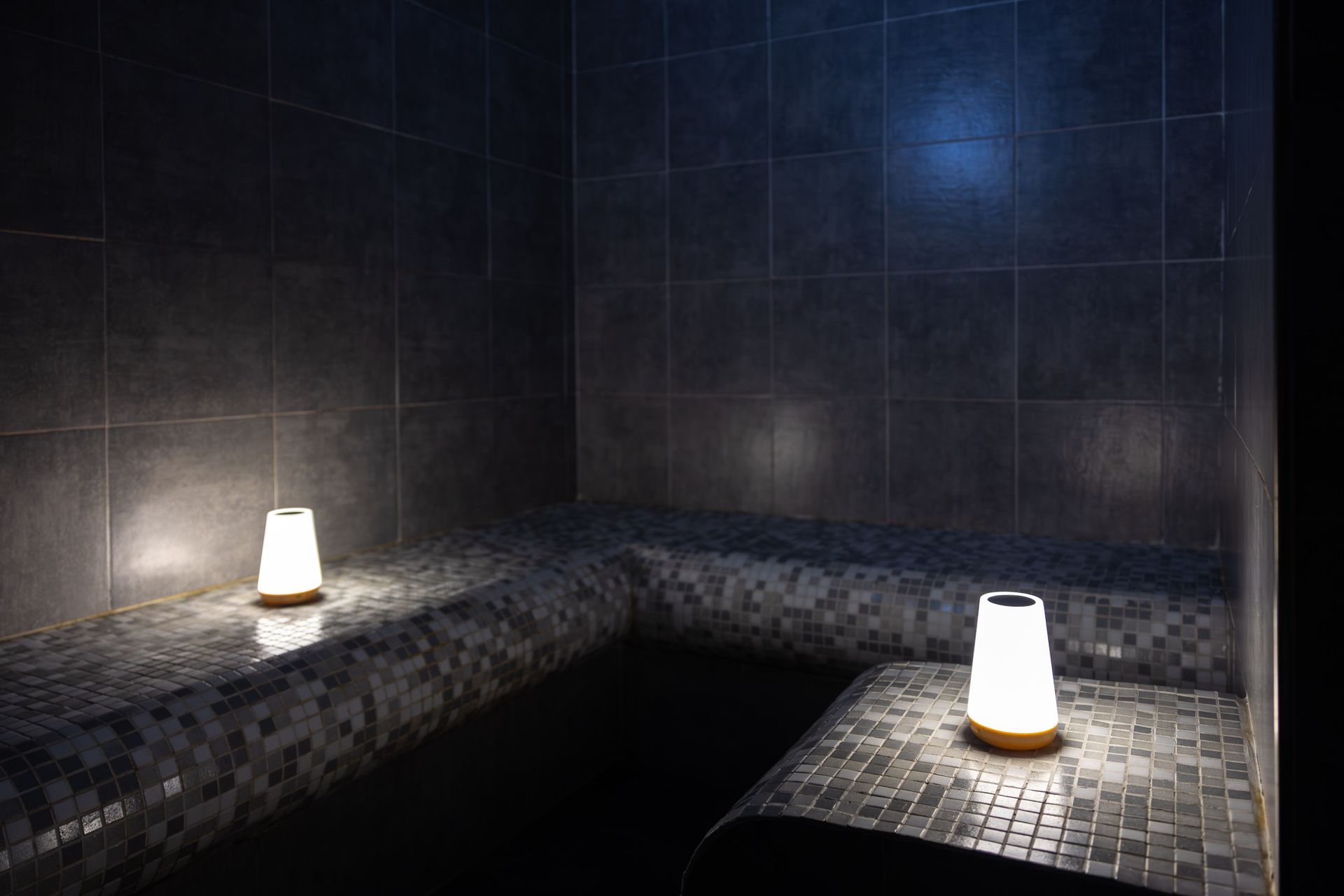 Stanza della spa in penombra con panche in mosaico scuro e due lampade luminose sulle mensole.