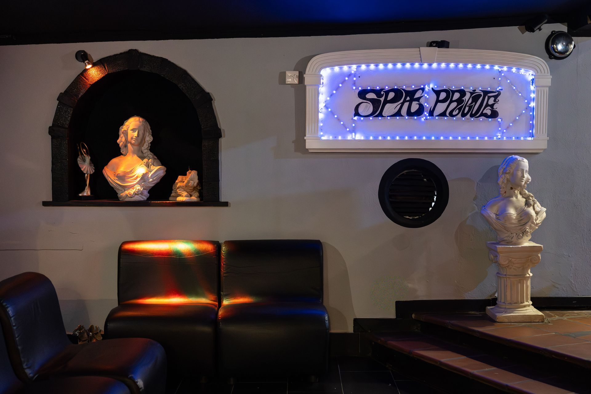 Salotto in penombra con insegna al neon, sculture a busto e poltrone in pelle scura su una teca a parete.