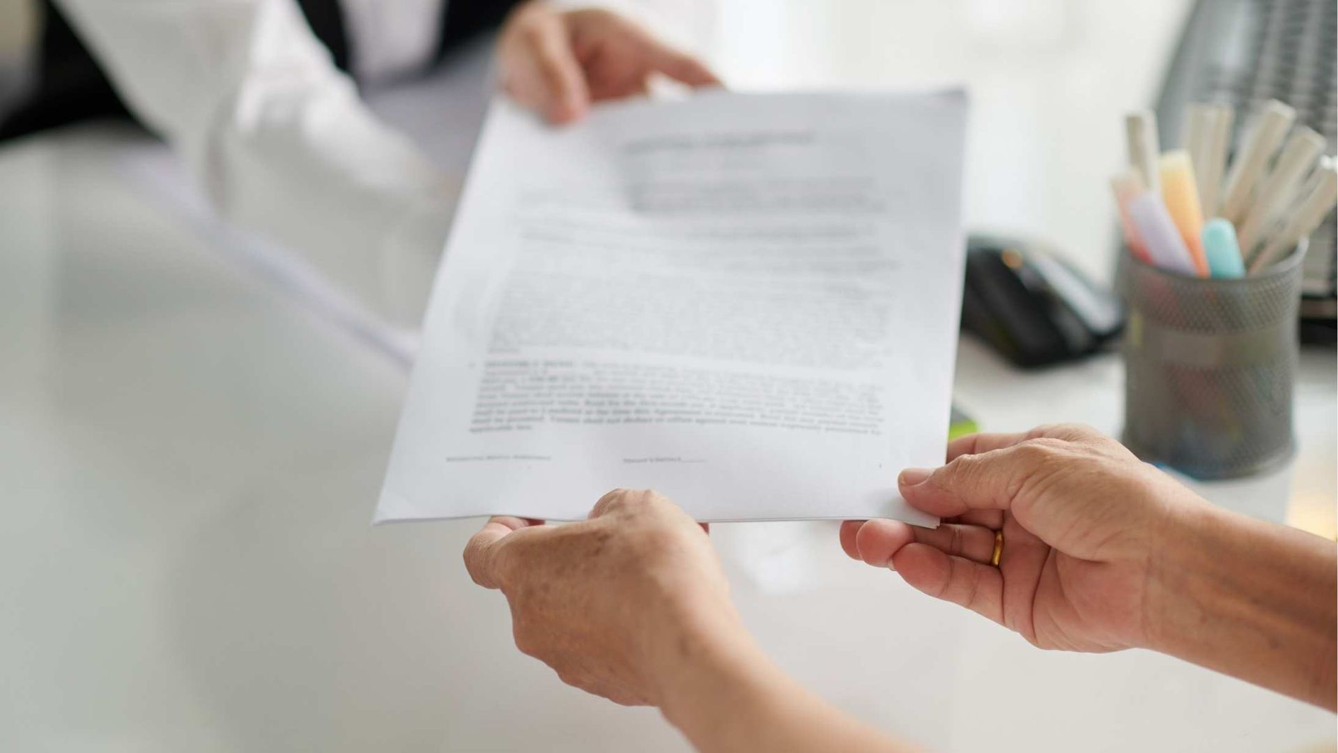 Hands exchanging a document in a bright office, with a pencil cup on the desk.