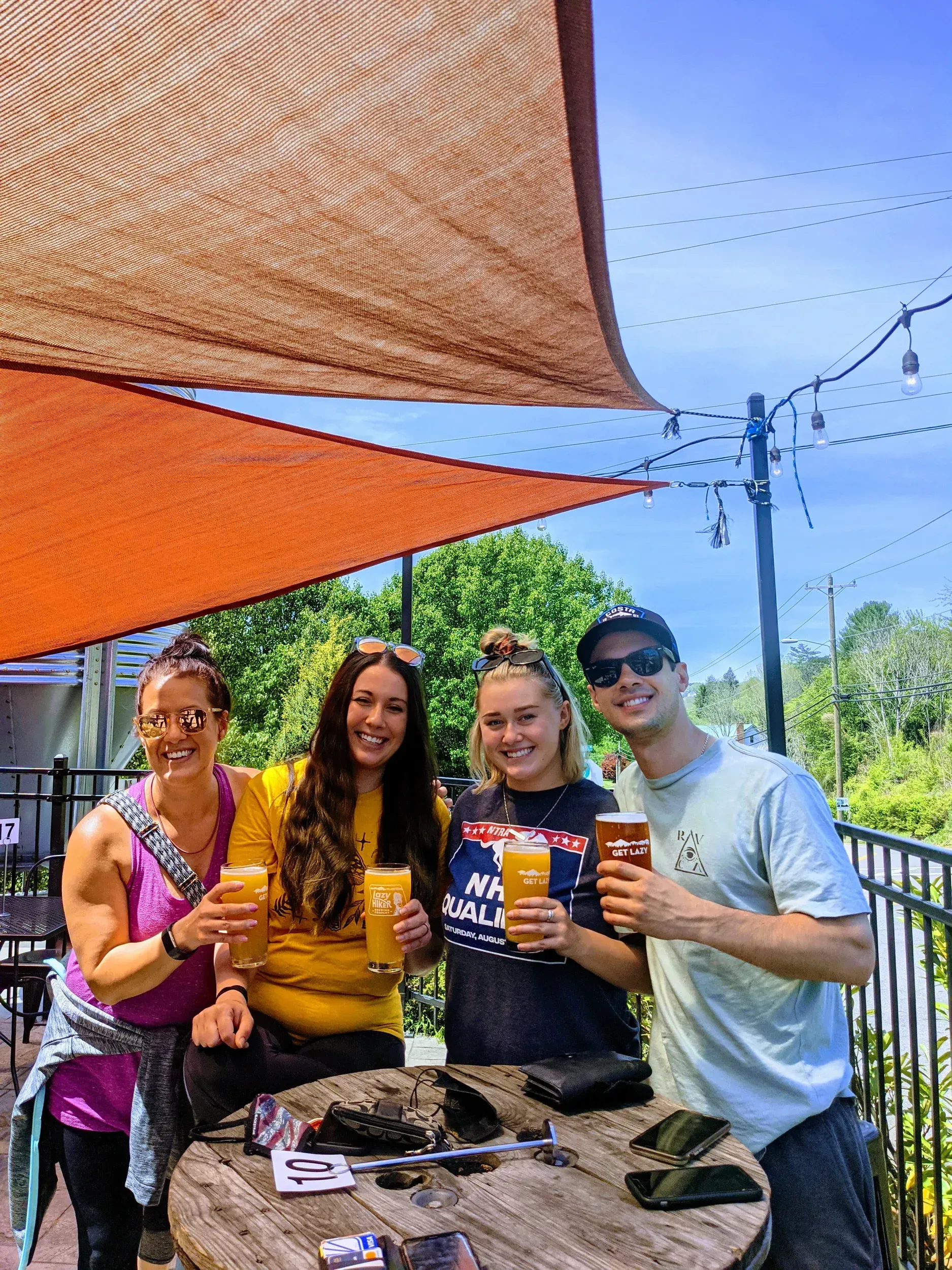 Four friends holding drinks and smiling under orange shade sails at an outdoor gathering