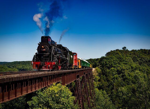 Steam locomotive crossing a high trestle bridge through a lush green valley under a blue sky