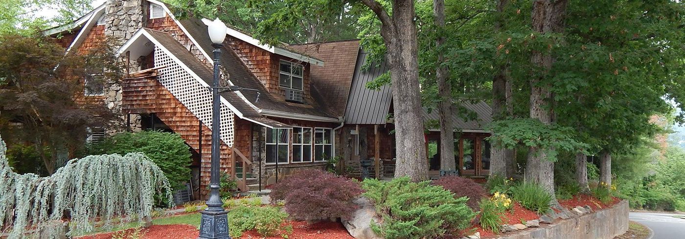 Brick house with white trim and red roof surrounded by tall trees and a landscaped yard