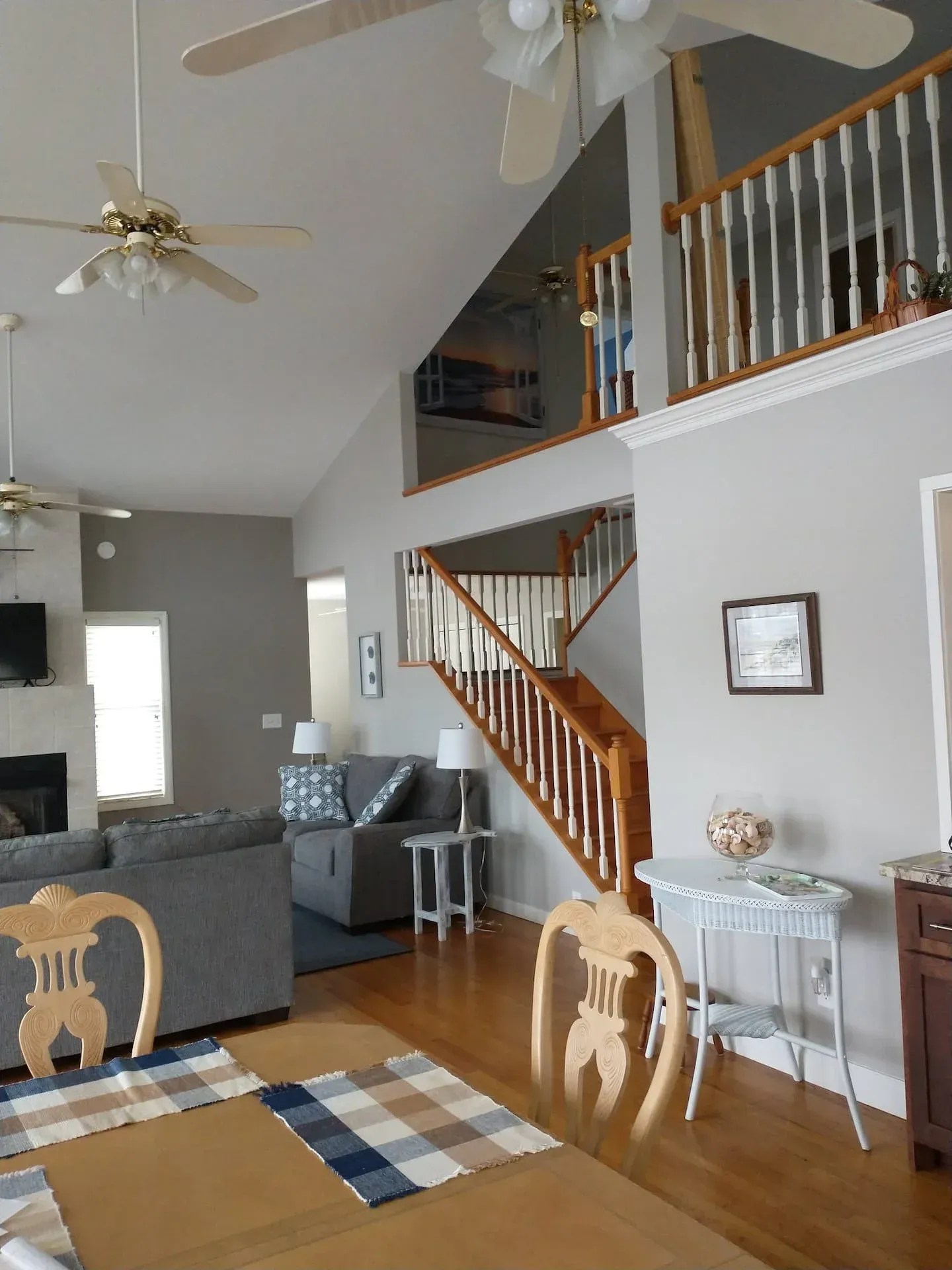 Living room with high ceiling, staircase, gray sofa, ceiling fans, and dining table in foreground