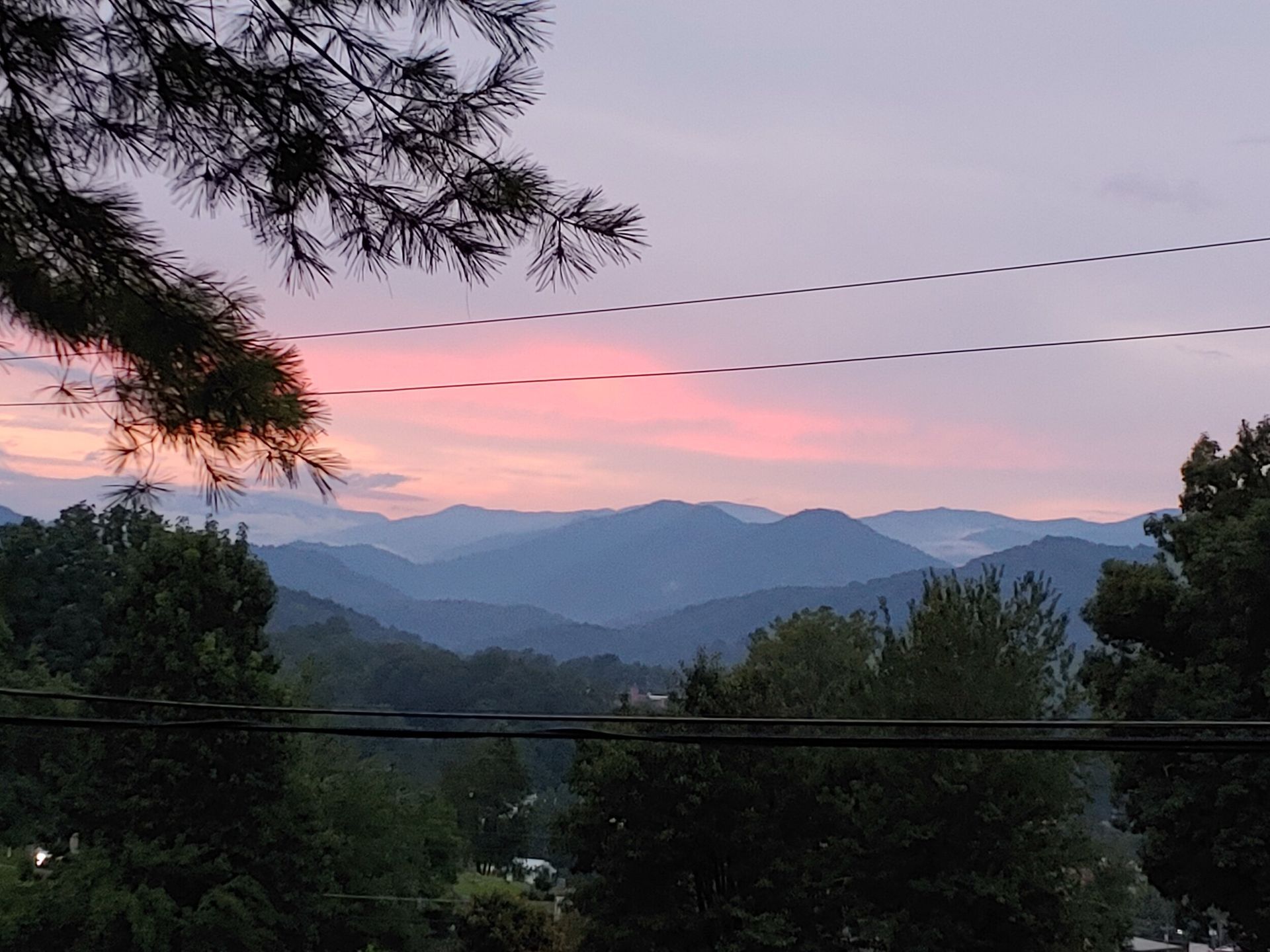 Pink sunset over layered mountains, framed by tree silhouettes and power lines.
