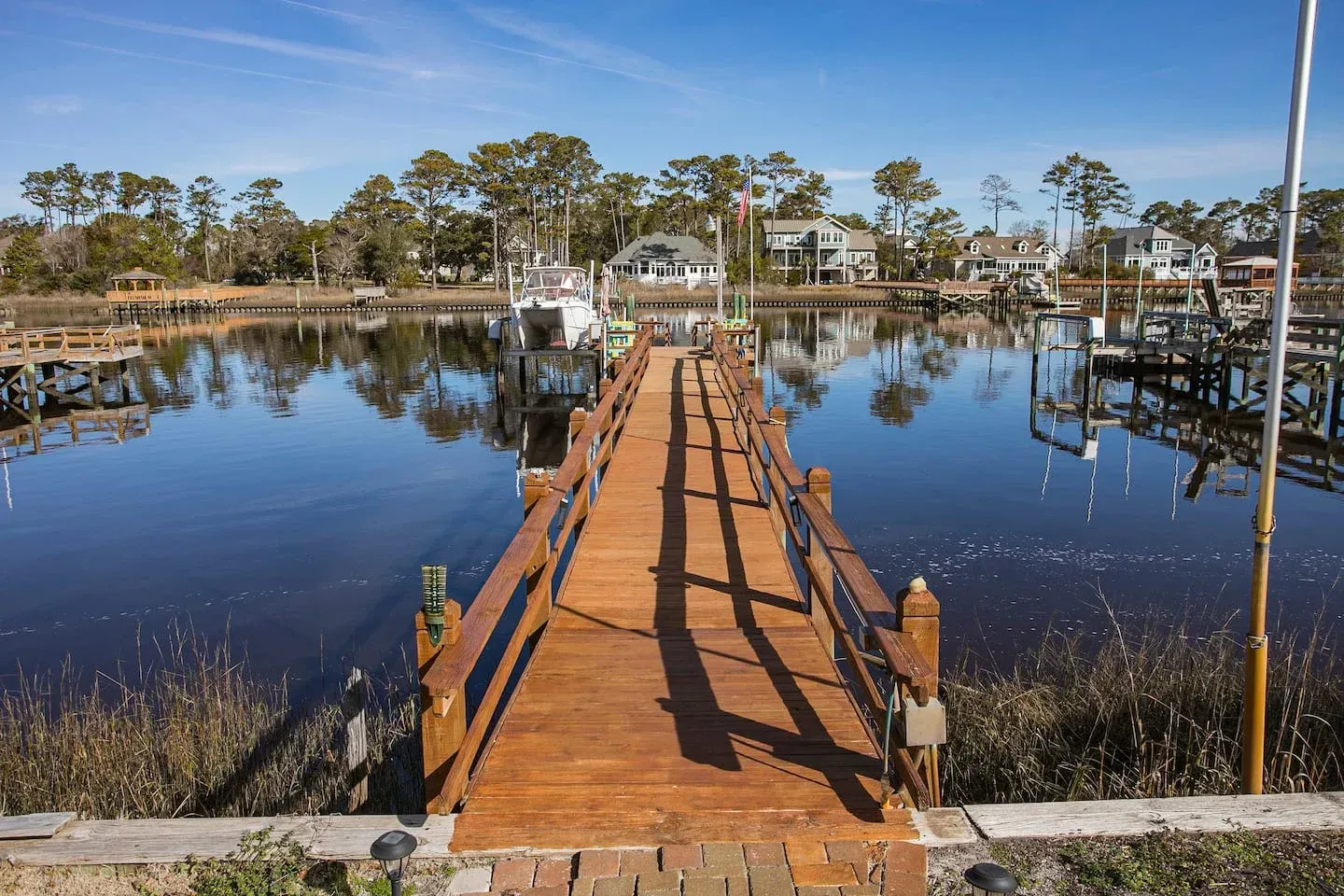 Wooden dock leading across a calm marina with boats, trees, and waterfront houses under a clear sky