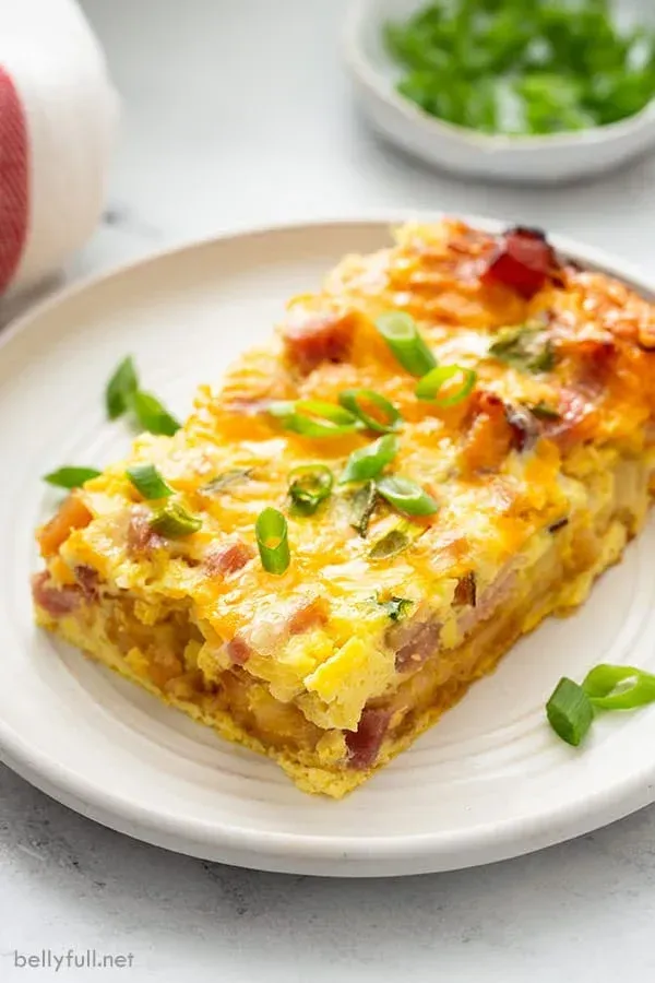 Cheesy baked egg casserole topped with green onions on a white plate, with a bowl in the background