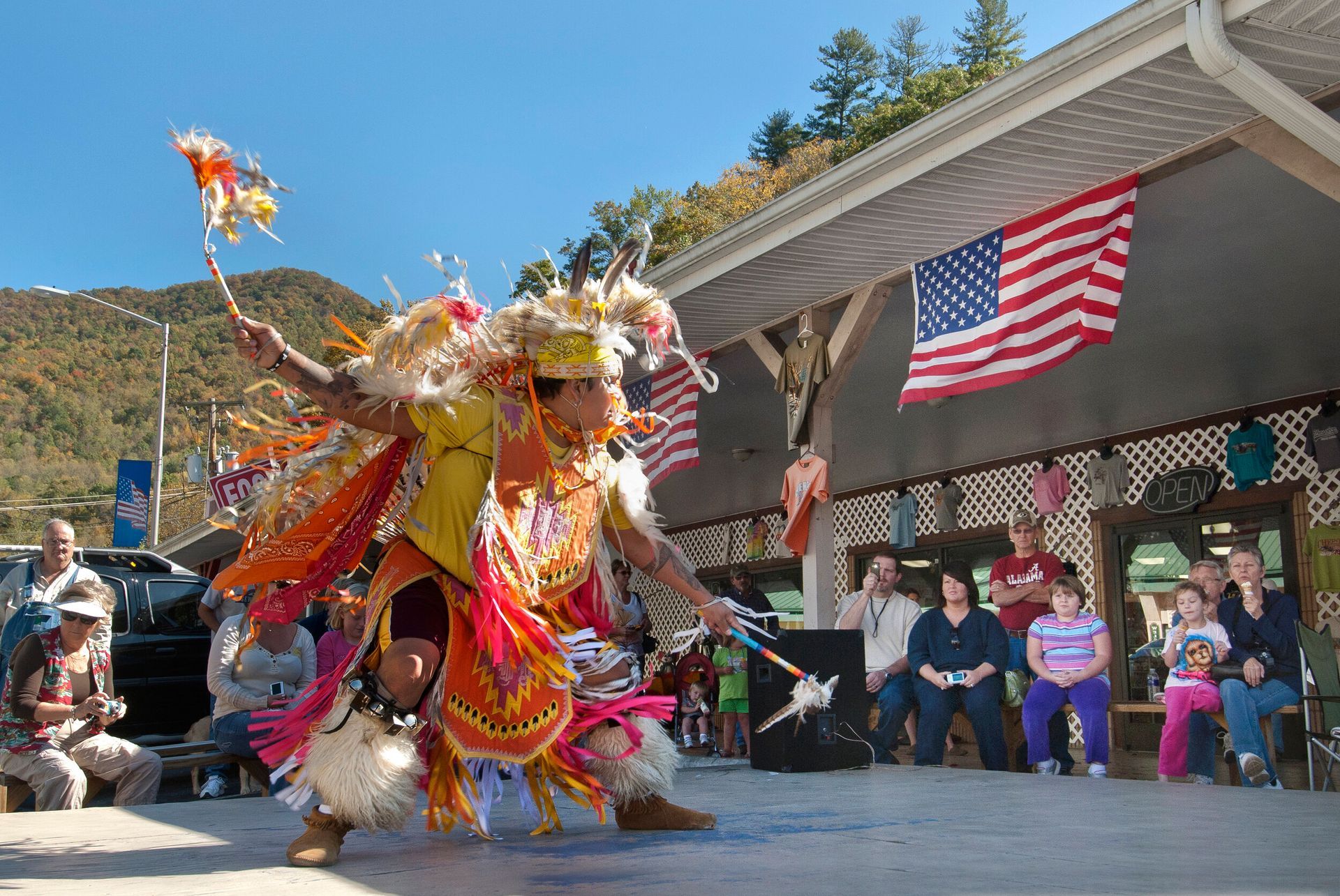 Colorful dancer performing outdoors in front of a building with an American flag and seated spectators