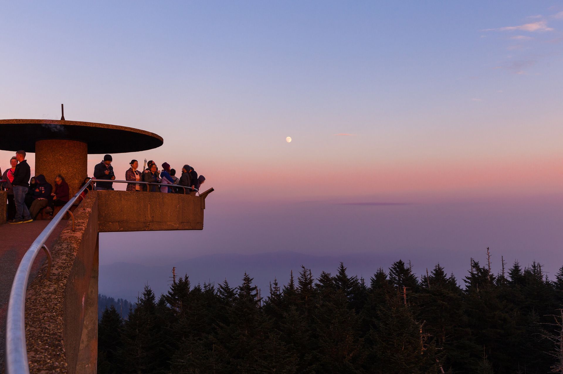 People gathered on a curved overlook at sunset above a forest, with a pink-purple sky.