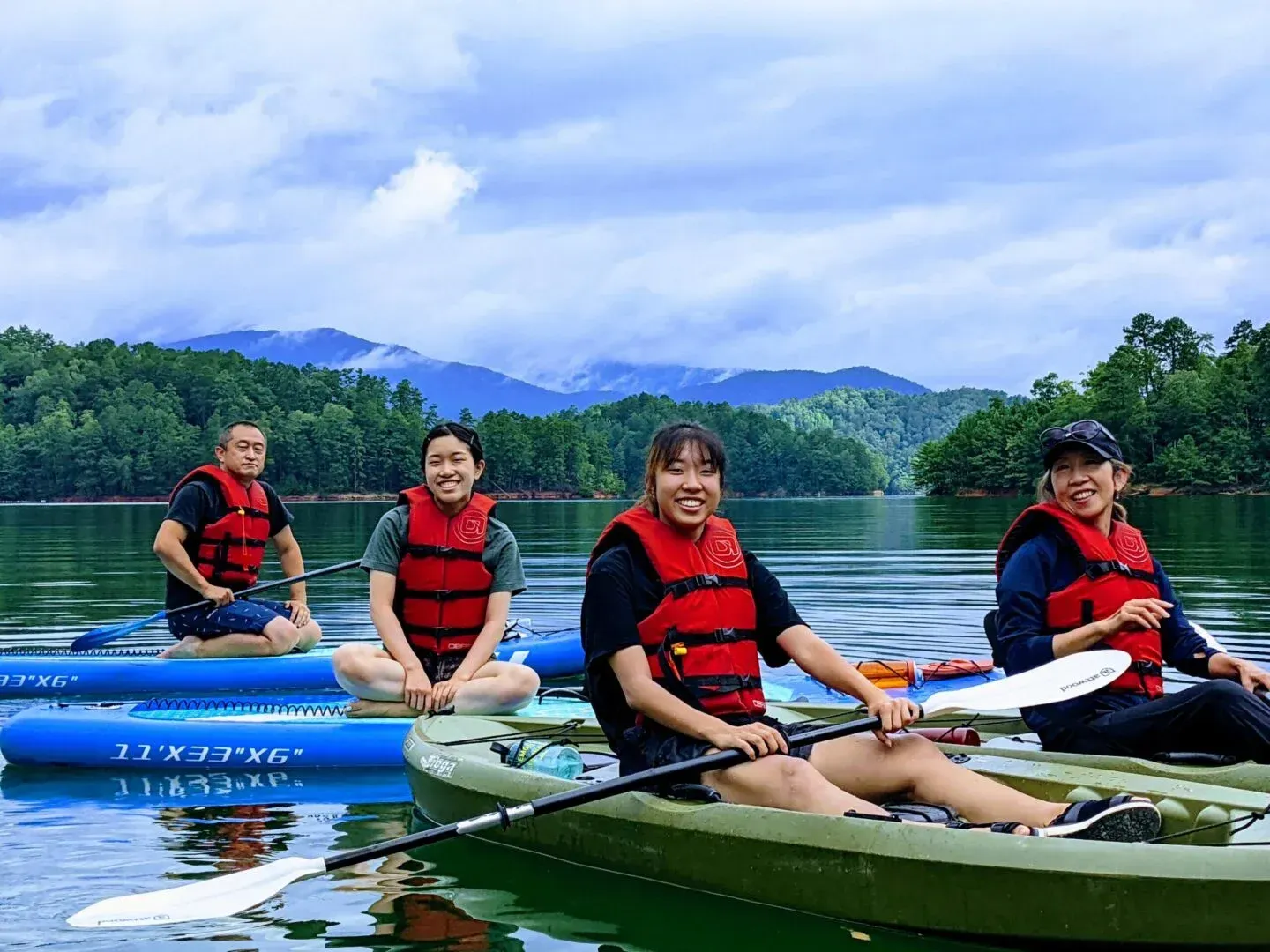 Four people in life vests kayak on a calm lake with mountains and cloudy sky behind them