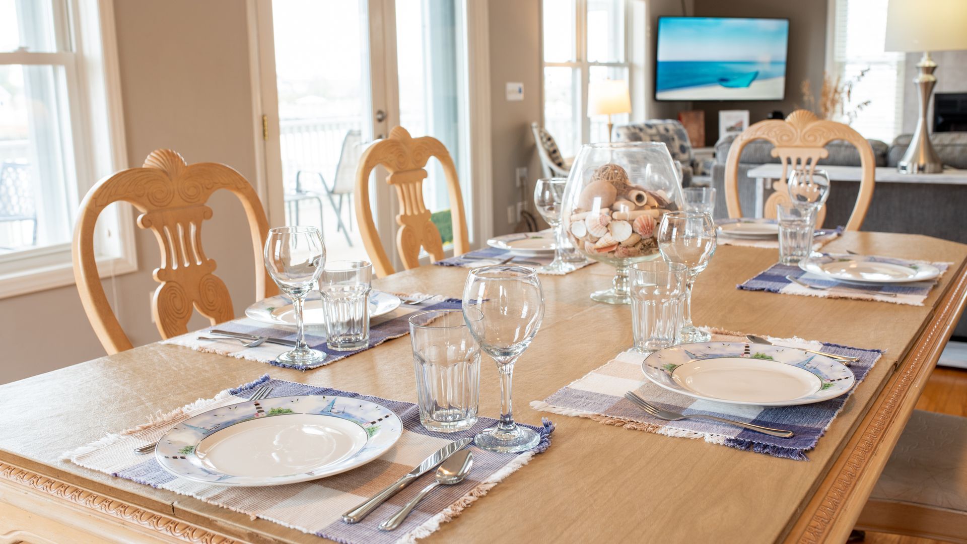 Set dining table in a bright room with white plates, glassware, and a seaside view on TV.