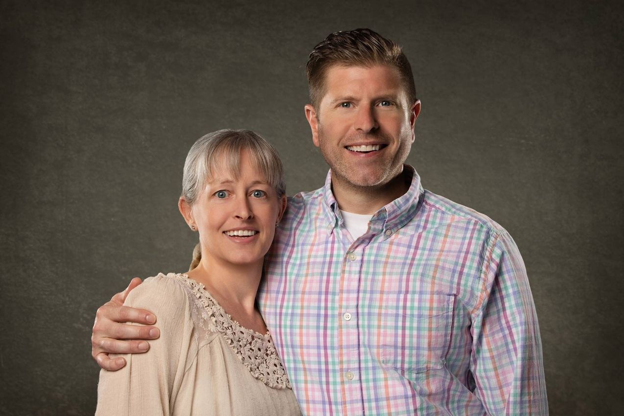 Two smiling people posing together against a gray background, one with an arm around the other