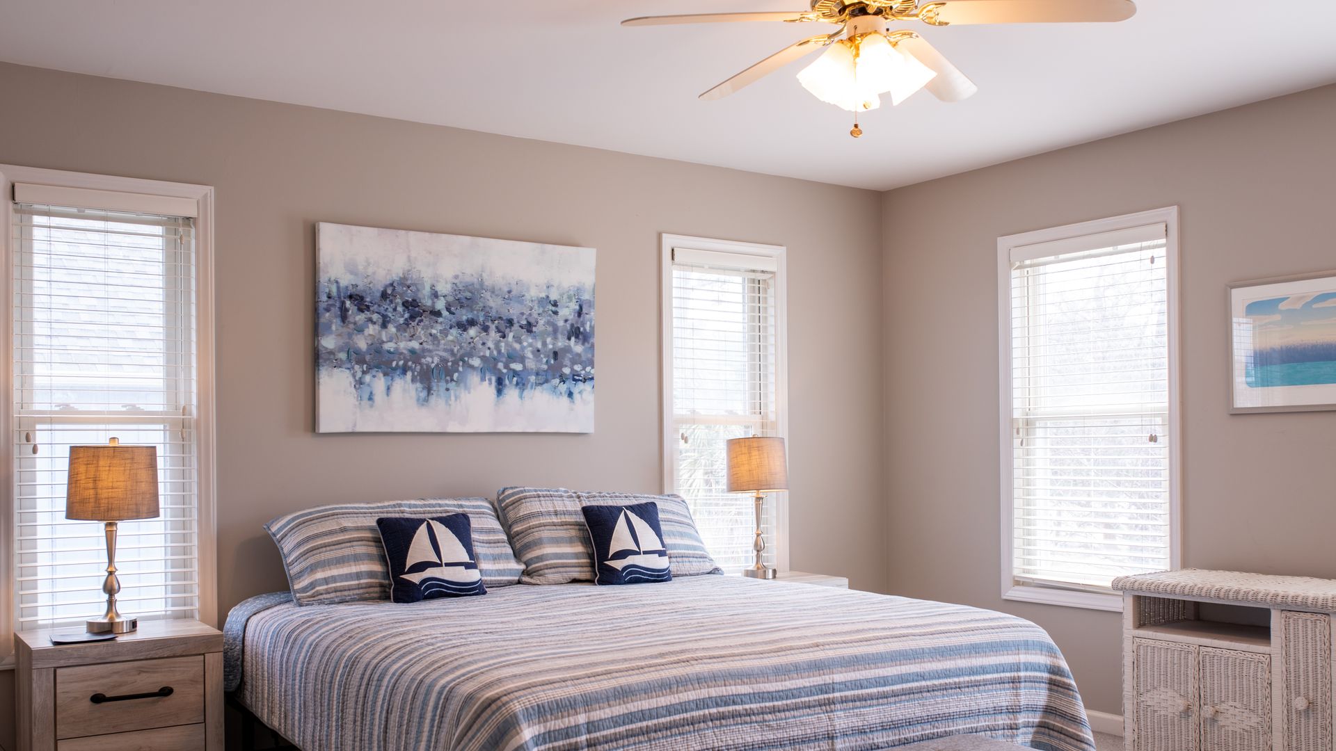 Bright bedroom with a blue striped bedspread, framed art, nightstands, and windows with blinds.