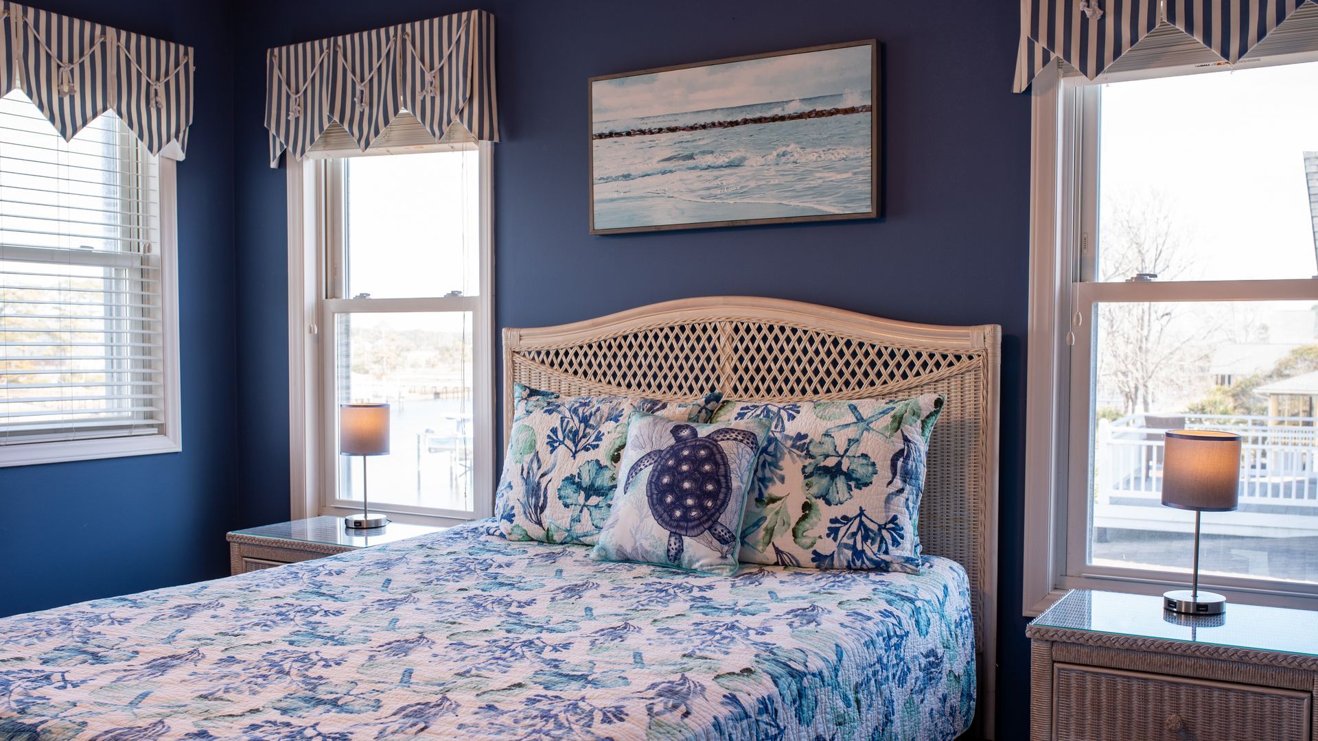 Blue bedroom with floral bedding, woven headboard, striped window valances, and coastal wall art