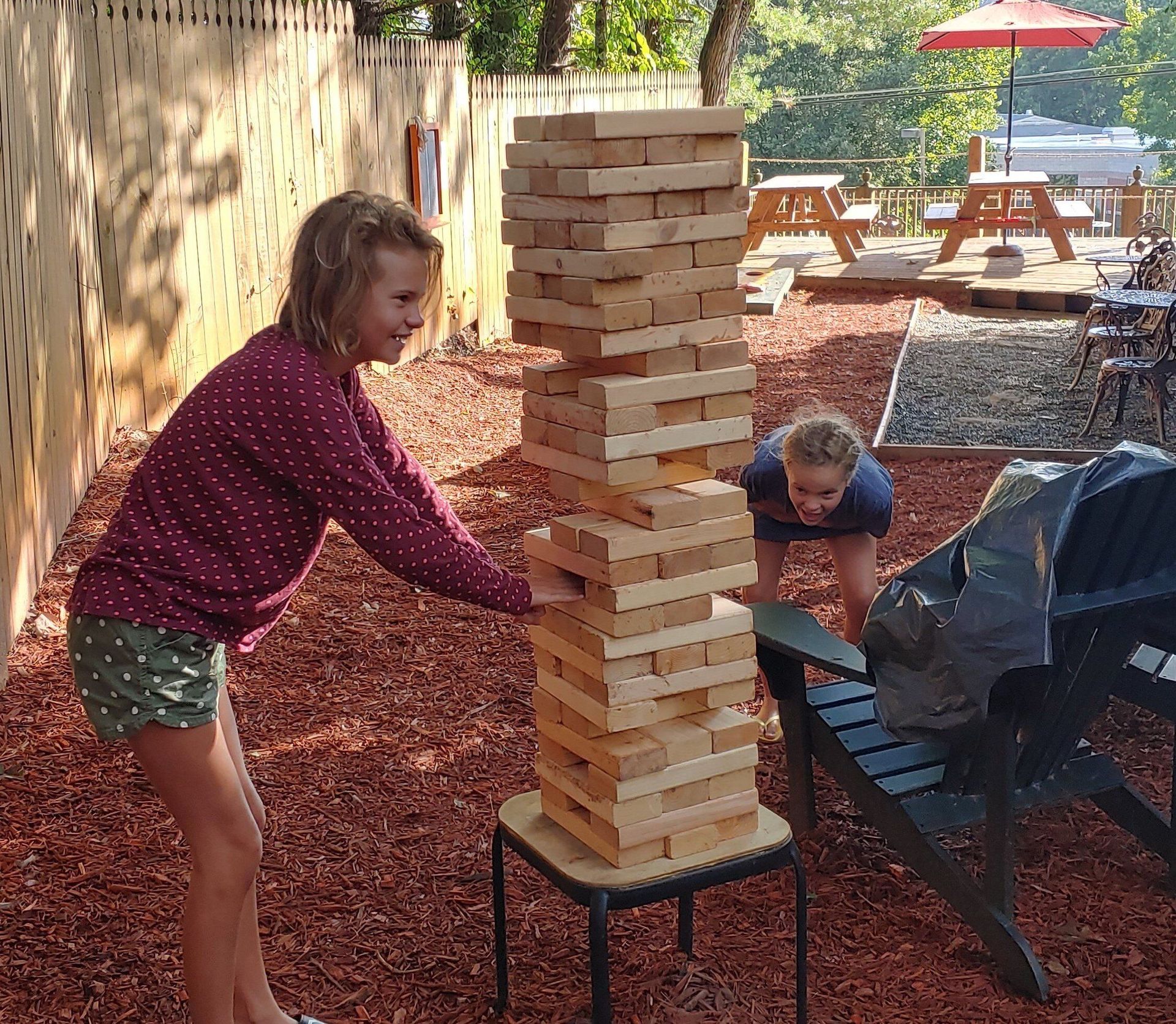 Two children play giant Jenga outdoors on a patio with red wood chips and a fence backdrop.