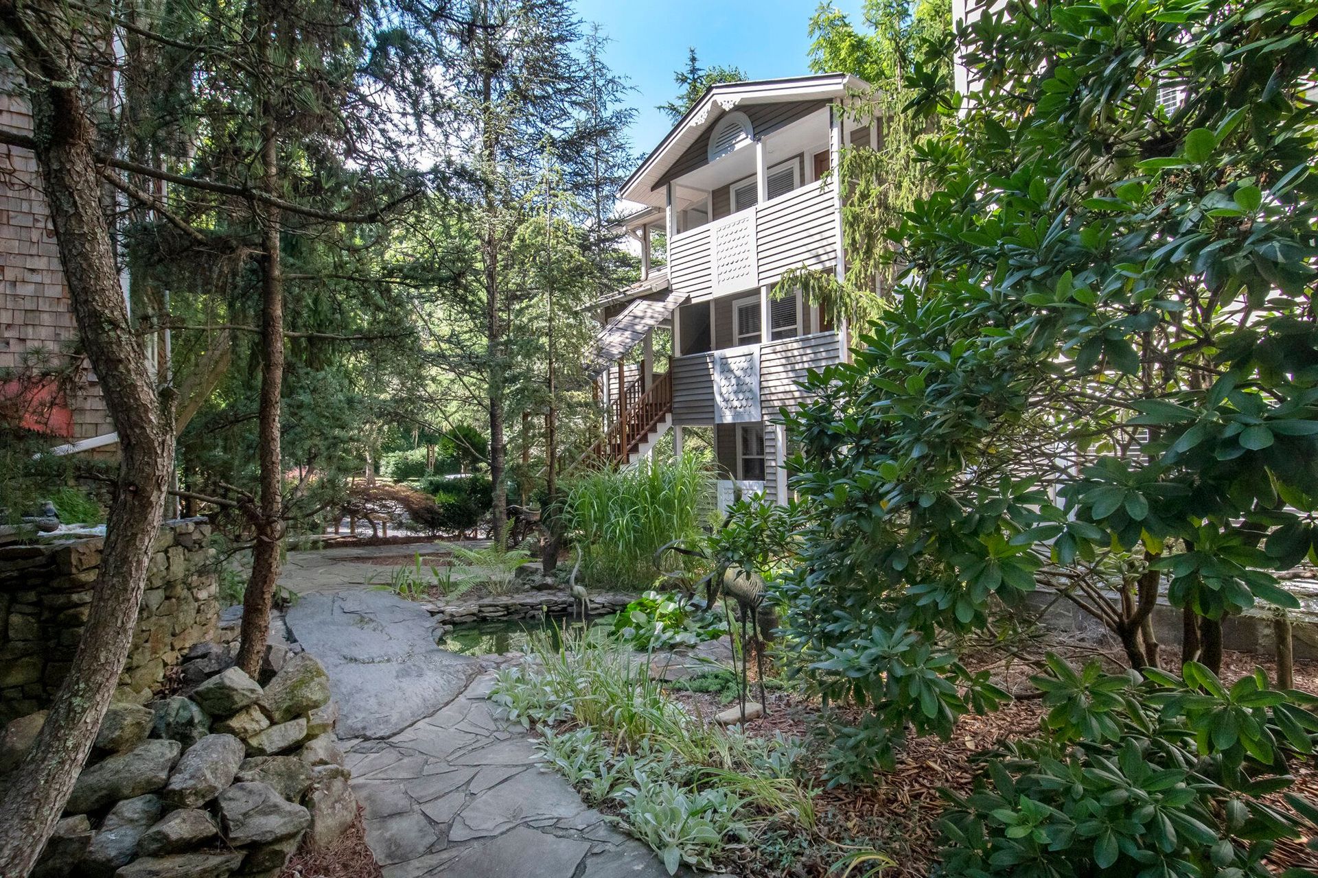 Forest path leading to a multi-story house with balconies, surrounded by trees and greenery